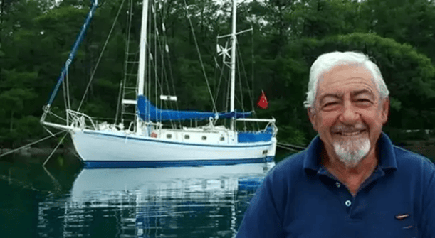 Bir adam gülümseyerek bir sailboat'un önünde duruyor, arka planda yeşil ağaçlarla çevrili su var.