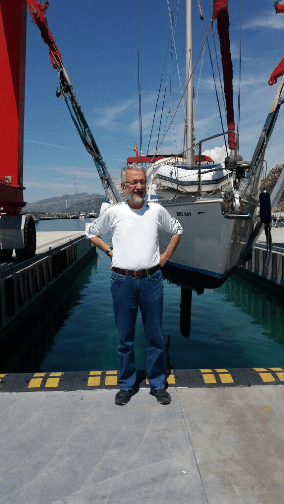 Bir iskelede, arka planda bir tekneyle birlikte poz veren beyaz tişörtlü bir adam.