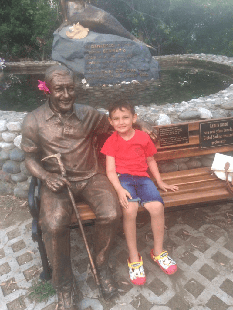 Bir çocuk, bronz bir heykelin yanında oturuyor. Heykel, Sadun Bora'yı temsil ediyor. Arka planda yeşil bir alan ve su kaynağı görülüyor.