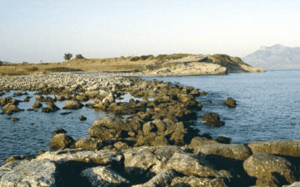 Sakin bir deniz manzarası, taşlarla kaplı kıyı ve uzak dağlar.